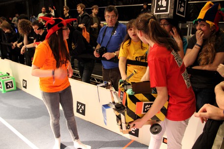 Two competitors standing in an arena, one holding a robot, talking to a team mate outside the arena