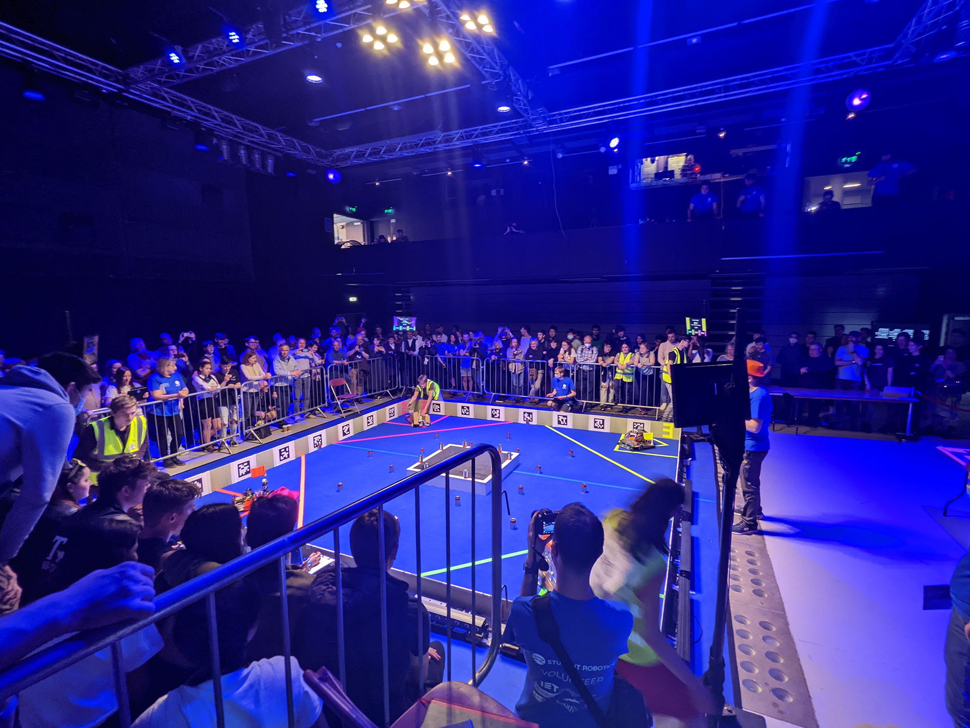 A crowd of onlookers
           looking into an arena with four robots, one in each corner