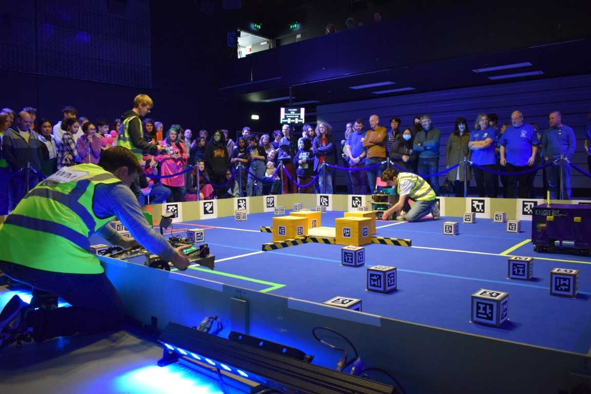 Three competitors placing their robots into the arena with a crowd of onlookers watching